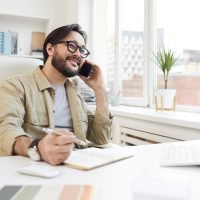 Communicative man calling customer Cheerful communicative young Asian man with beard sitting at table in office and calling customer on phone